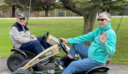 Two seniors driving four-wheeled peddle cars and smiling.