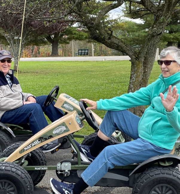 Two seniors driving four-wheeled peddle cars and smiling.